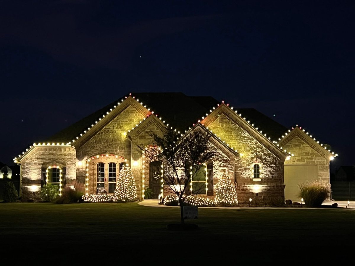 Stone home with warm white roofline lighting and lit shrubs across the front yard
